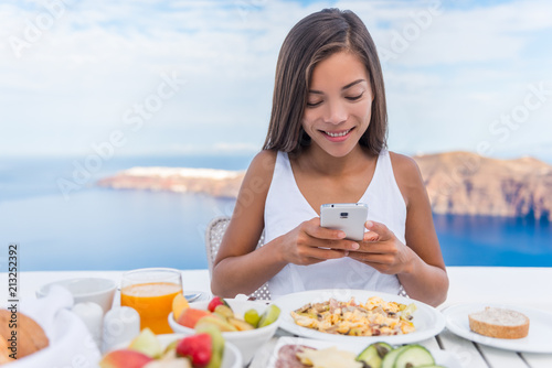 Food selfie social media vlogging girl taking photo with phone of breakfast plate using cellphone app. Luxury travel vacation lifestyle. Beautiful model in resort in Santorini, Greece, Europe. © Maridav