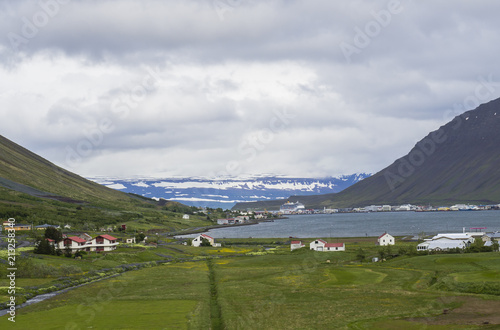 Beautiful view on Isafjordur city  capital of west fjords of Iceland in summer with red houses  harbor with ships and yachts  green grass and farm landscape  blue snow covered mountains background
