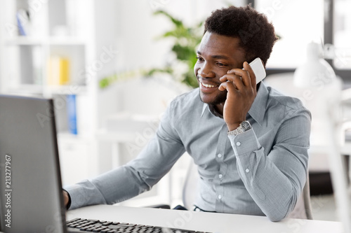 business, people, communication and technology concept - smiling african american businessman with computer calling on smartphone at office