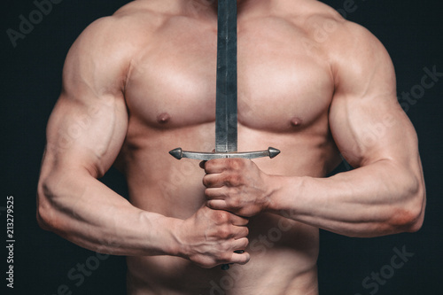 Bodybuilder man posing with a sword isolated on black background. Serious shirtless man demonstrating his mascular body photo