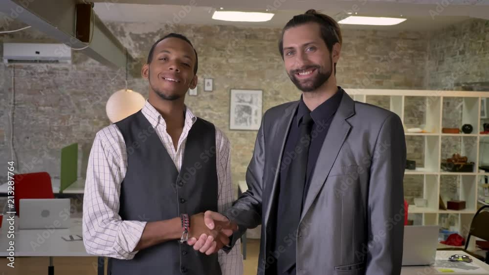 Two multy-ethnic workers are shaking hands in office, watching at camera, smiling, communication concept Two multy-ethnic workers are shaking hands in office, watching at camera, smiling, communication concept