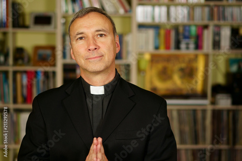 A good looking catholic priest is praying into his library. He looks at us with serenity, optimism, joy, admiration, trust, confident and peace.