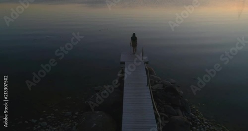 Woman on a wharf, C4K aerial descending tilt view a woman, standing at a wooden dock, enjoying the sun and a warm summer sunset, on a sunny evening dusk, in Hanko, Uusimaa, Finland