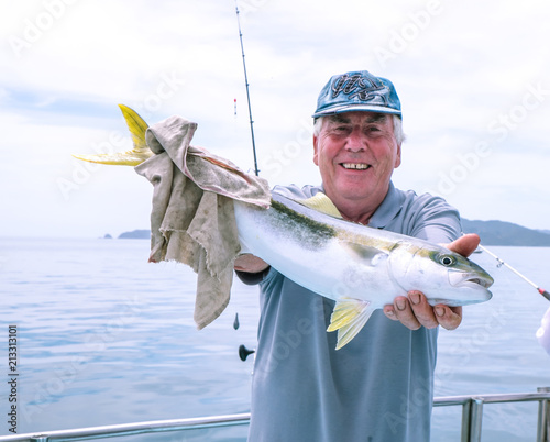 Happy mature senior caucasian tourist has caught a kingfish on a fishing charter boat at Doubtless Bay, Far North, Northland, New Zealand, NZ