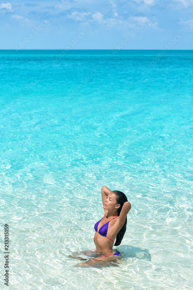 Bikini body beach swimwear model swimming in idyllic turquoise ocean water relaxing under the sun. Asian woman touching her healthy hair in swimsuit tanning body.