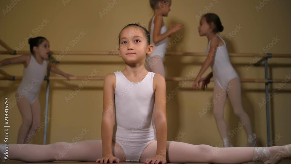 Little Asian girl in a white swimsuit and pink skirt shows ballet exercises in the foreground with other girls in the background in a ballet studio. Stock ビデオ | Adobe Stock