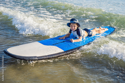 Happy baby boy - young surfer ride on surfboard with fun on sea waves. Active family lifestyle, kids outdoor water sport lessons and swimming activity in surf camp. Summer vacation with child photo