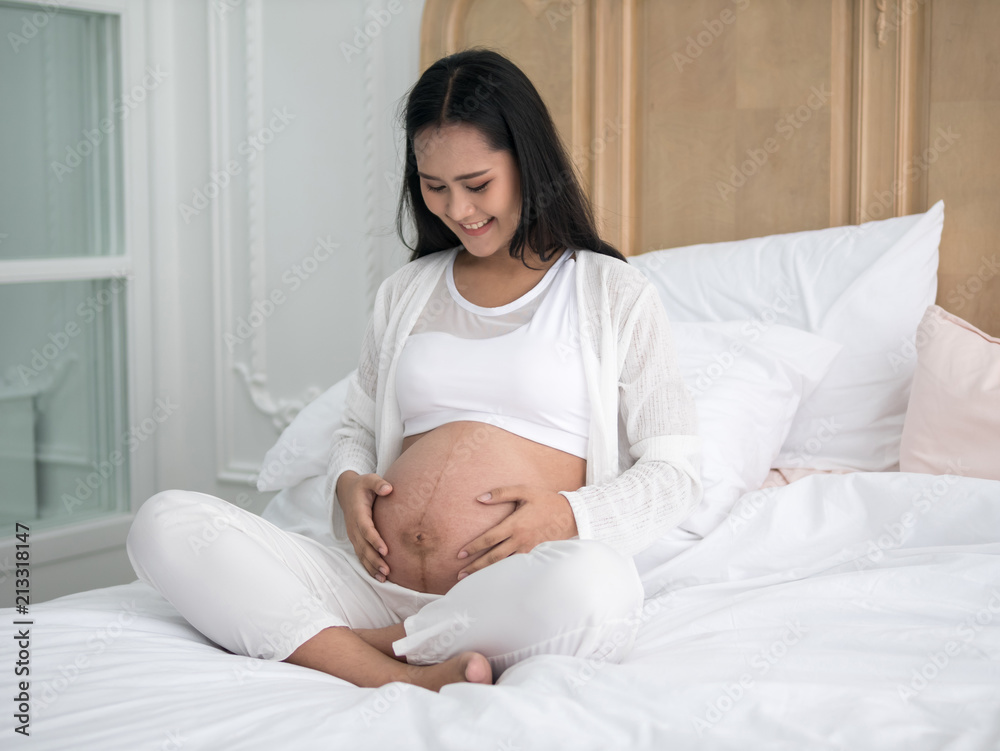 Happy pregnant asian woman sitting on bed and touching her belly in bedroom at home Happy pregnant asian woman sitting on bed and touching her belly in bedroom at home