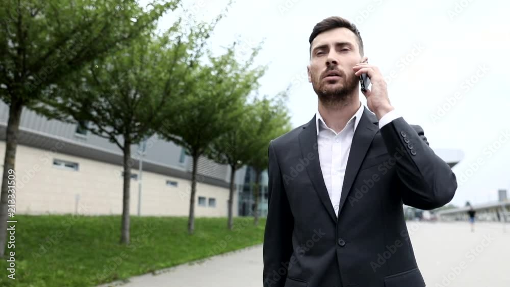 Close up of Attractive Businessman Walking in the City. Talking on his Modern Smartphone. Wearing Official Black Suit. Successful life. Business style. Confident Gait. Close up of Attractive Businessman Walking in the City. Talking on his Modern Smartphone. Wearing Official Black Suit. Successful life. Business style. Confident Gait.