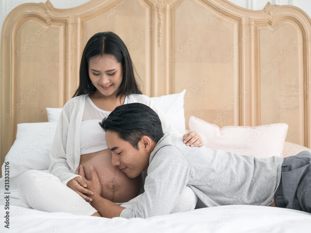 Fototapeta premium Beautiful asian pregnant woman and her handsome husband laying on the bed while spending time together. Man listening to baby in belly. Happy family lifestyle.