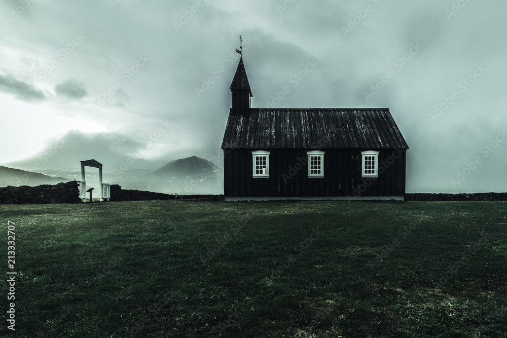 a black church with white window frames and a gate infront of the church taken from the side on a misty rainy windy dark moody day in Iceland