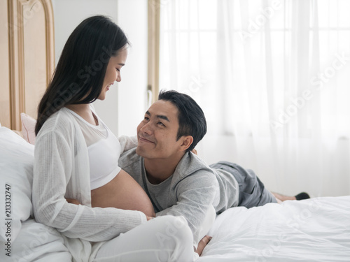 Beautiful asian pregnant woman and her handsome husband laying on the bed while spending time together. Man listening to baby in belly. Happy family lifestyle.