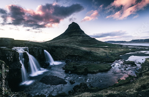 Kirkjufell Mountain in Iceland in the background with three waterfalls in silky look in the front with a river flowing around a mountain into the ocean and a colorful pink and purple sky with clouds