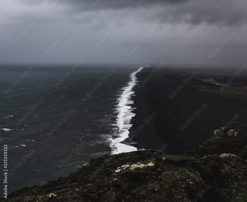 Bird perspective on a coastline in Iceland with a black sand beach with black and white contrasts between the waves and the beach while a storm is approaching with dark clouds in the sky