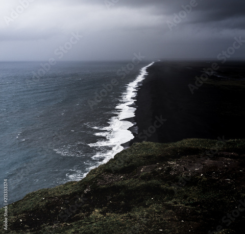 Bird perspective on a coastline in Iceland with a black sand beach with black and white contrasts between the waves and the beach while a storm is approaching with dark clouds in the sky