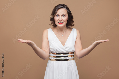 Portrait of angry middle aged boss woman looking at camera. Emotional expressing woman in white dress, red lips and dark curly hairstyle. Studio shot, isolated on beige or light brown background