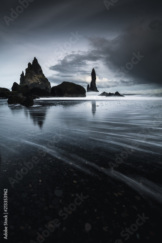 Rocks on a black sand beach in Iceland with reflection in the Sea and a dark sky in misty moody weather with dark colors and a rough sea in landscape format with a storm approaching