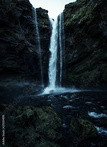 A wild natural fresh sparkling waterfall in a lonely green valley with moos covered rocks and ice cold mountain water flowing down black rocky walls on a rainy moody day in Iceland without tourists