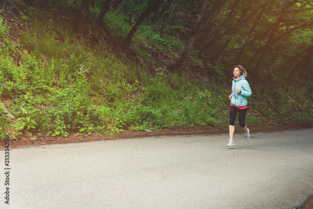 A young blonde woman running is practicing outdoors in a city mountain park in the forest. Warm rays through the branches of trees