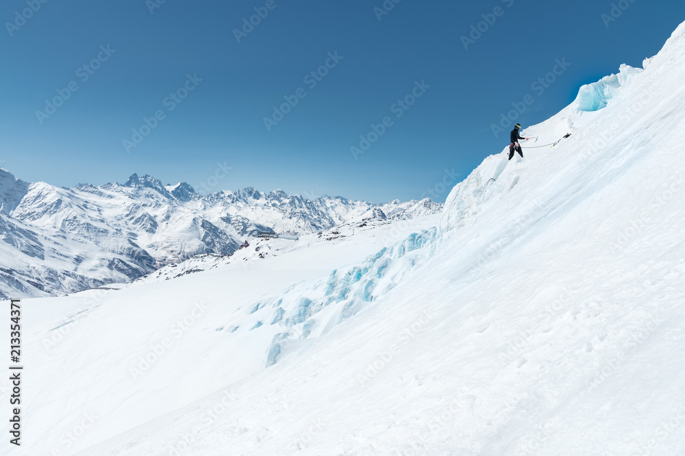 A professional mountaineer in a helmet and ski mask on insurance makes a nick-hole in the glacier against the backdrop of the Caucasian snow-capped mountains