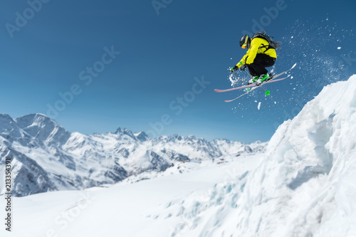 A skier in full sports equipment jumps into the precipice from the top of the glacier against the background of the blue sky and the Caucasian snow-capped mountains. Elbrus region. Russia