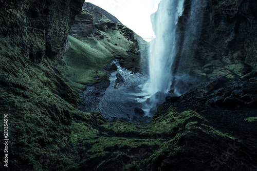 A wild natural fresh sparkling waterfall in a lonely green valley with moos covered rocks and ice cold mountain water flowing down black rocky walls on a rainy moody day in Iceland without tourists