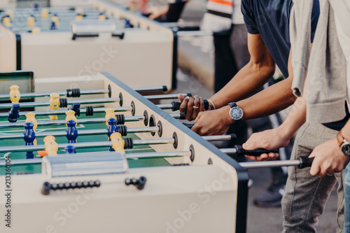 Two men stand on one side, play table football together, enjoy heat games, like kicker, spend free time outdoor. Team and companionship concept