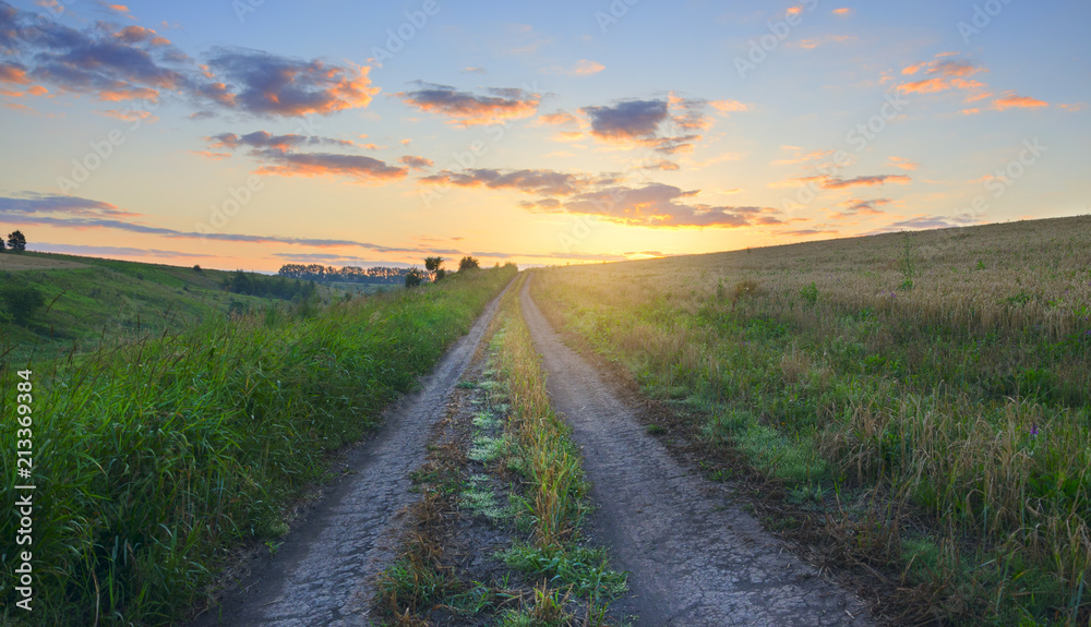 Beautiful summer scene with ground country road on a background of rising sun and colorful sky and clouds.