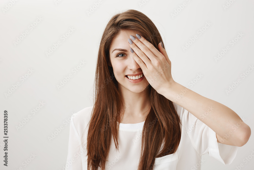 My eyes see everything. Studio shot of sincere pretty caucasian brunette with white smile, covering one eye while standing against gray background, checking vision in optician shop My eyes see everything. Studio shot of sincere pretty caucasian brunette with white smile, covering one eye while standing against gray background, checking vision in optician shop