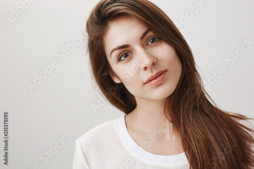 One can drown in her eyes. Portrait of gorgeous natural brunette tilting head while listening with interest, charmed by speaker, gazing at camera with desire and curiosity, standing over gray wall
