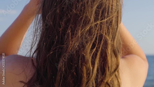 Caucasian young woman playing with her long brown hair in sunlight on a beach looking at the sea, back view, slow motion photo