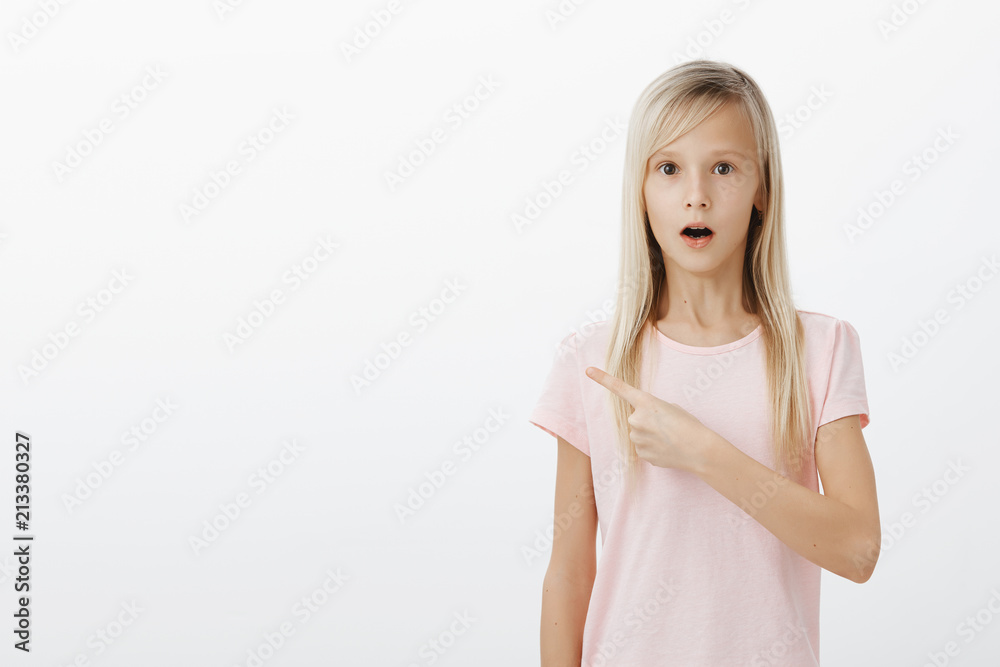 Girl showing classmate shocking sign on blackboard. Indoor shot of amazed surprised little child with fair hair, dropping jaw and gasping from wonder, pointing at upper left corner over gray wall Girl showing classmate shocking sign on blackboard. Indoor shot of amazed surprised little child with fair hair, dropping jaw and gasping from wonder, pointing at upper left corner over gray wall