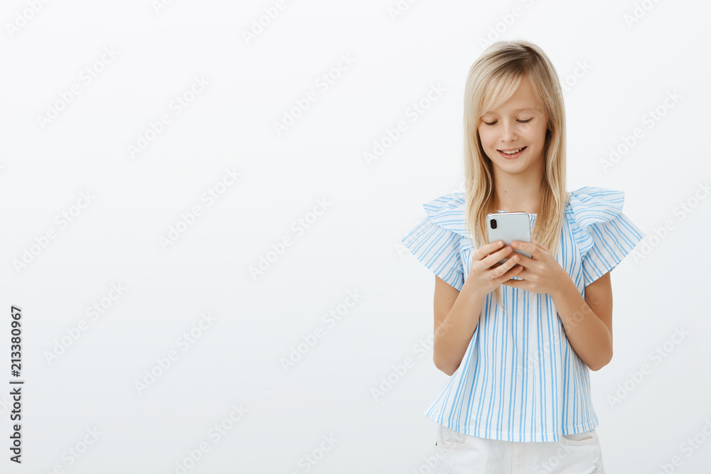 Studio shot of pleased good-looking small female kid with blond hair in blue blouse, holding smartphone and smiling at screen while messaging or playing funny game over gray background Studio shot of pleased good-looking small female kid with blond hair in blue blouse, holding smartphone and smiling at screen while messaging or playing funny game over gray background