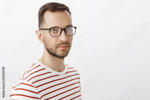 Studio shot of serious handsome bearded man in black glasses, looking aside with casual unemotive expression, standing over gray background, posing for advertisement or waiting for friend near house © Cookie Studio