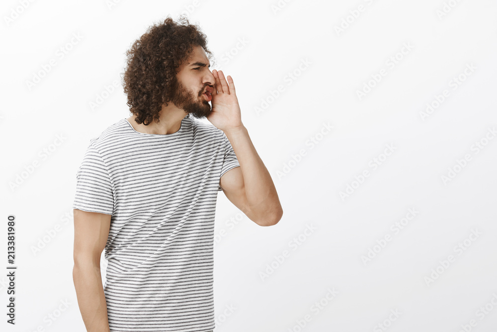 Portrait of pissed off impolite hispanic guy, turning right and holding palm near mouth while shouting cursing words out loud, frowning, being bothered and angry, standing displeased over gray wall Portrait of pissed off impolite hispanic guy, turning right and holding palm near mouth while shouting cursing words out loud, frowning, being bothered and angry, standing displeased over gray wall