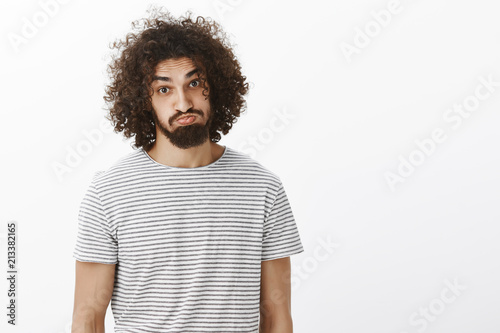 Portrait of plyaful carefree attractive male model with beard and curly hair, sticking out tongue and pouting, making funny faces while hanging out on beach with friends, standing over gray background