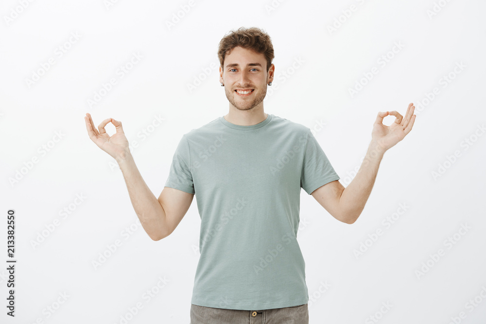Pleased relaxed handsome guy in earrings and casual t-shirt, smiling broadly, standing against gray background with spread hands in zen gesture and meditating or being patient while practicing yoga Pleased relaxed handsome guy in earrings and casual t-shirt, smiling broadly, standing against gray background with spread hands in zen gesture and meditating or being patient while practicing yoga