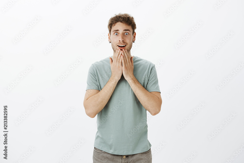 Studio shot of excited upbeat attractive male in earrings and t-shirt, holding palms on jaw and gasping, starring with popped eyes with disbelief, being impressed and amazed with best gift ever Studio shot of excited upbeat attractive male in earrings and t-shirt, holding palms on jaw and gasping, starring with popped eyes with disbelief, being impressed and amazed with best gift ever