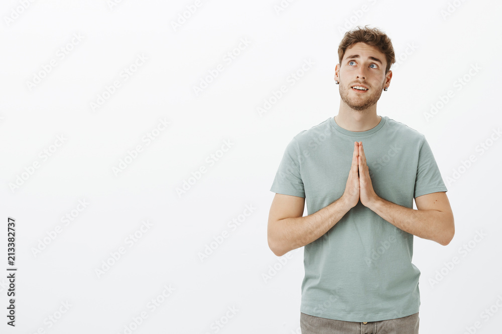 Indoor shot of dreamy hopeful attractive guy in t-shirt, holding hands in pray and looking up with thoughtful expression, praying or hoping, making wish for better life, posing over gray background Indoor shot of dreamy hopeful attractive guy in t-shirt, holding hands in pray and looking up with thoughtful expression, praying or hoping, making wish for better life, posing over gray background