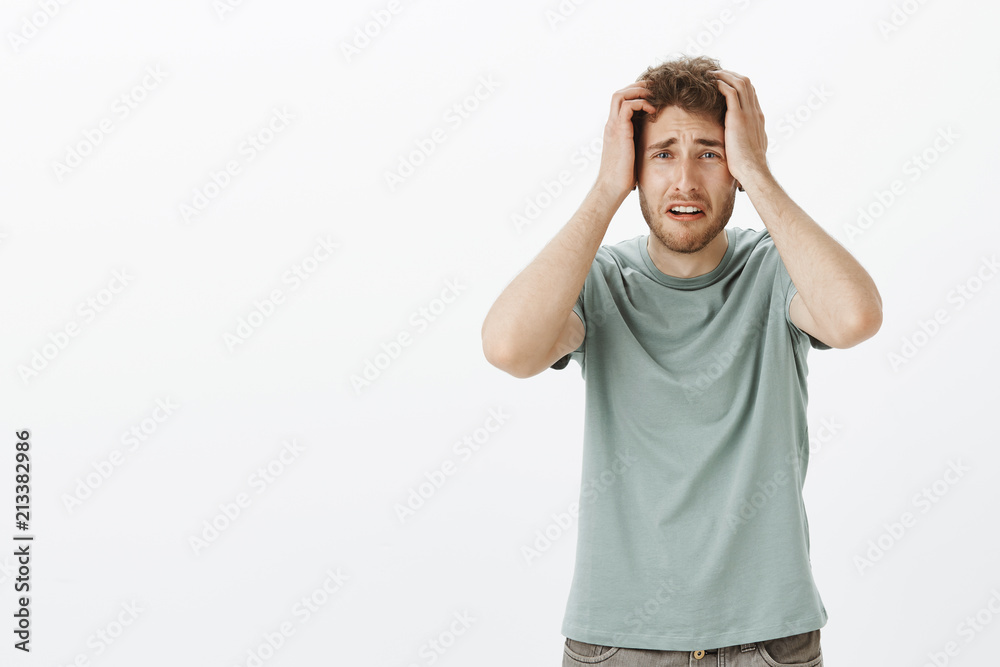 Fototapeta premium Portrait of troubled devasted young male who lost job, holding hands on head and frowning, feeling worried and anxious while seeing terrible accident, standing upset over gray background