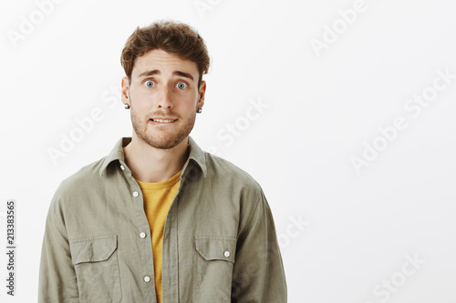 Guy making silly face wanting girlfriend forgive him. Charming bearded male in trendy shirt, biting lip and frowning, admitting fault, feeling remorse, standing over gray background