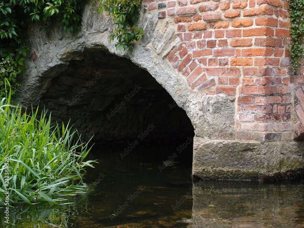 A brick built tunnel channels a river under a European urban center on a summer day.