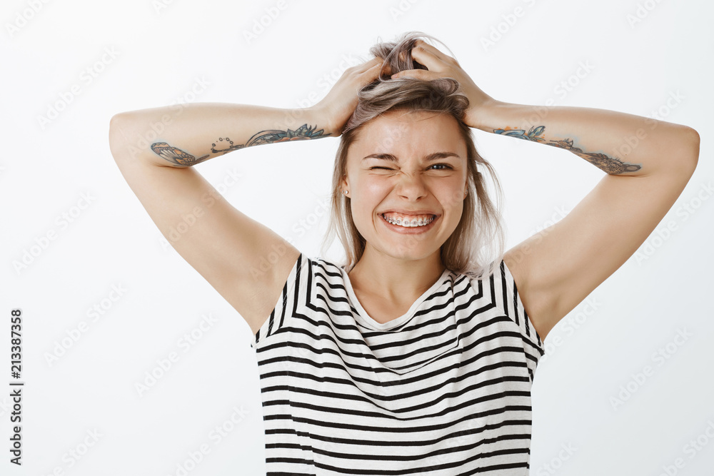 Good-looking emotive girlfriend with fair hair and tattooed arms, holding hands on head and smiling broadly showing cute braces, winking while being in great mood, amused over gray background