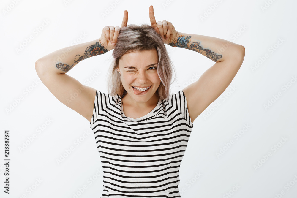 Stubborn good-looking girlfriend with fair hair and tattoos, winking and smiling broadly, showing cute braces on teeth, making horns from index fingers on head, being playful and flirty