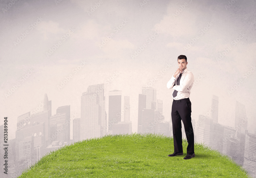 A confident male well looking office manager standing in small green grass in front of city landscape with tall buildings concept. A confident male well looking office manager standing in small green grass in front of city landscape with tall buildings concept.