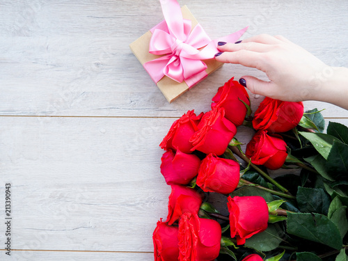 Preparing a gift, on a wooden table, with roses. Female hand holding a gift, wrapped with ribbon, on a background of red roses, the space under the text photo