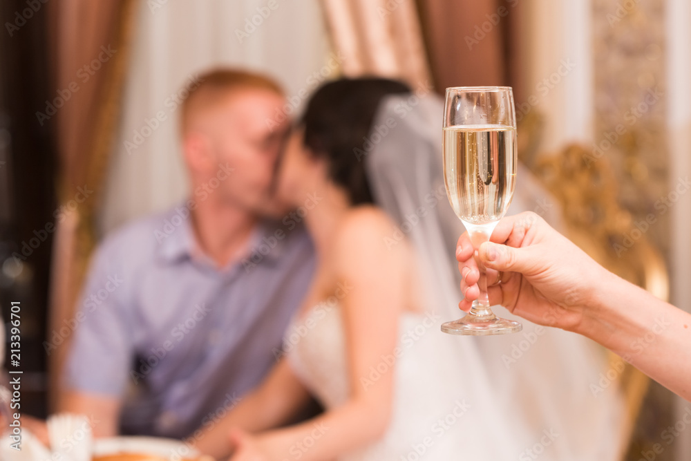 A glass of champagne in a woman's hand against the background of kissing newlyweds in the blur