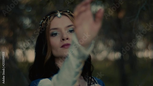 Brunette woman in image of medieval witch is waving hand in front of camera. Close-up of her face in a forest, she is looking calmly