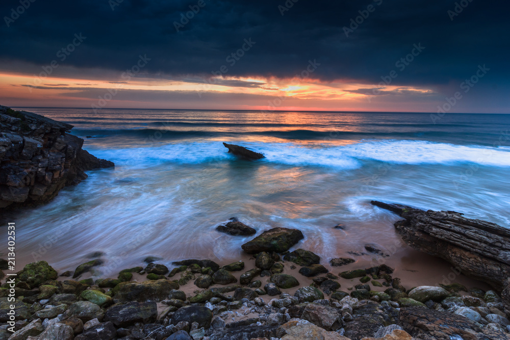 Fototapeta premium Amazing sunset on the ocean. View of dramatic cloudy sky and stony coastal rocks. Atlantic coast near Lisbon. Portugal.