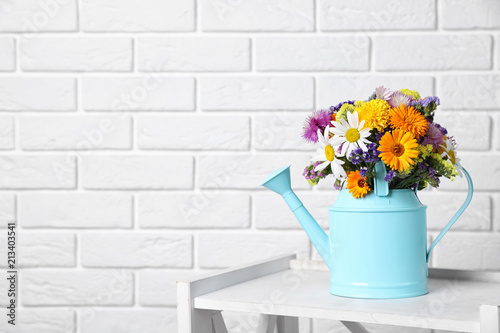 Watering can with beautiful wild flowers on table near brick wall